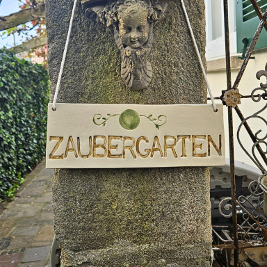 Schild mit Dekor Blume des Lebens und der Aufschrift "Zaubergarten" an einer Steinmauer.