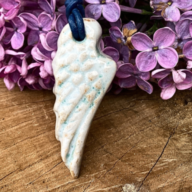 Ein kleiner, hellblauer Anhänger in Form eines Engelsflügels, umgeben von lila Blüten.