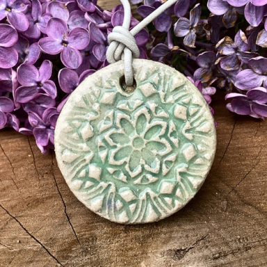 Runde, grüne Keramikmedaille mit floralem Muster, umgeben von lila Fliederblüten.
