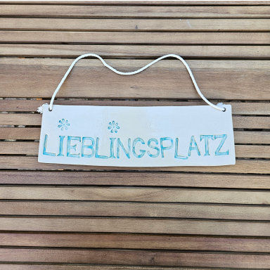Schild mit der Aufschrift „LIEBLINGSSPLATZ“ in blauer Schrift auf einem Holzuntergrund.