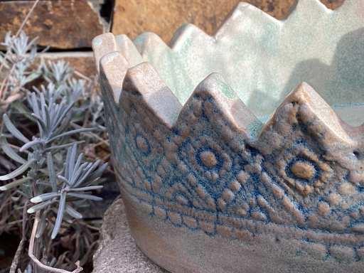 Dekorativer blauer Blumentopf mit zackigem Rand und geometrischem Muster, umgeben von Pflanzen.