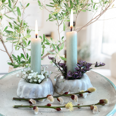 Zwei Kerzen in dekorativen Gugelhupf Kerzenständern, umgeben von frischen Blumen und Zweigen.