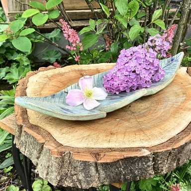 Keramik Schale, Federschale mit lila Blumen und einer Blüte auf einem Holzstamm vor grünen Pflanzen.