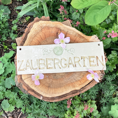 Schild mit der Aufschrift „Zaubergarten“ auf einem Holzstück, umgeben von Pflanzen.