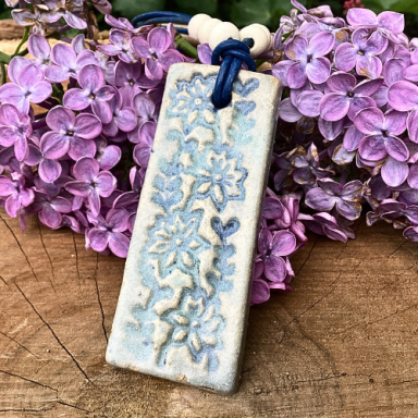 Blauer Anhänger mit floralen Mustern vor lila Fliederblüten.