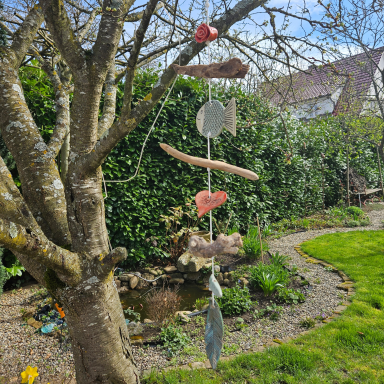 Ein Mobile aus Holz und Fischen hängt an einem Baum in einem Garten mit grünem Hintergrund.